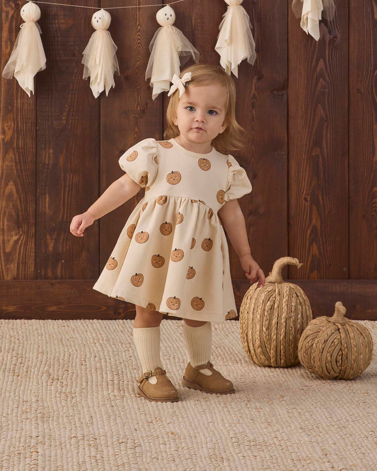 A toddler wearing the quincy mae darla dress in pumpkins—a cream fleece dress with a pumpkin print—stands on a beige rug near two textured pumpkin decorations, with a wooden wall and a hanging tassel garland in the background.