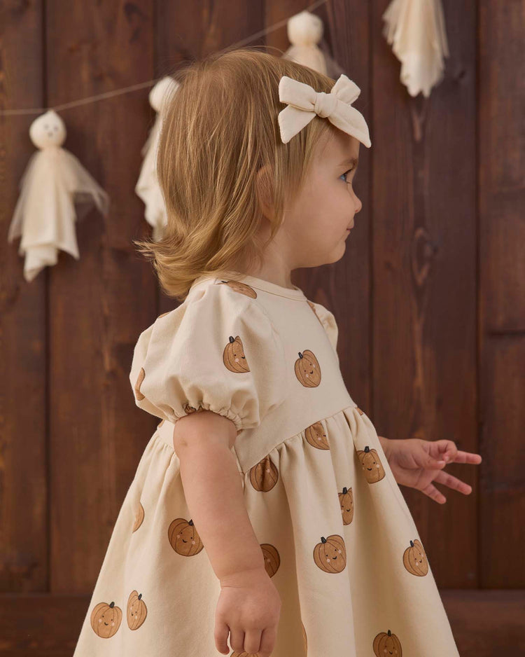 A young child wearing the quincy mae darla dress in pumpkins—a cream fleece dress with a cheerful pumpkin print and a matching bow—stands sideways against a wooden backdrop decorated with hanging ghosts.