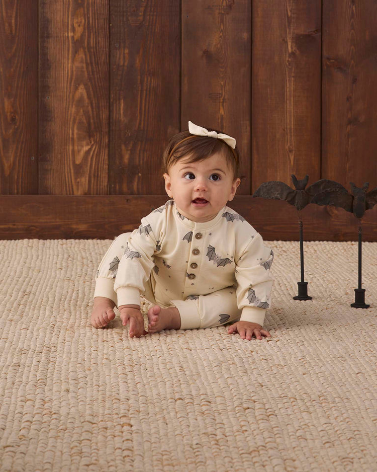 A baby wearing the quincy mae relaxed fleece jumpsuit in baby bats, made from soft organic cotton and featuring a cream bat print, with a white bow, sits on a textured rug in front of a wooden wall while two small black bat decorations rest nearby.