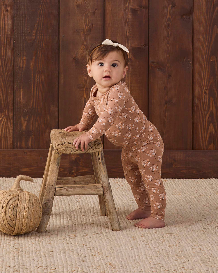 A baby wearing the quincy mae bamboo pajama set in fall floral stands holding a small wooden stool, with a ball of thick rope nearby, on a textured rug and wooden wall background.