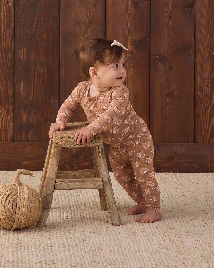 A baby wearing the Quincy Mae bamboo pajama set in fall floral stands holding a small wooden stool, looking to the side. The background features a wooden wall and a cream woven rug with a large ball of rope on the floor.