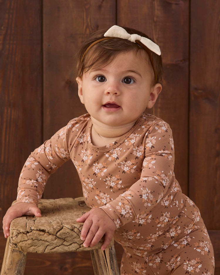 A baby wearing the quincy mae bamboo pajama set in fall floral stands holding onto a rustic wooden stool, with a dark wooden background behind, and is accessorized with a cream bow headband.