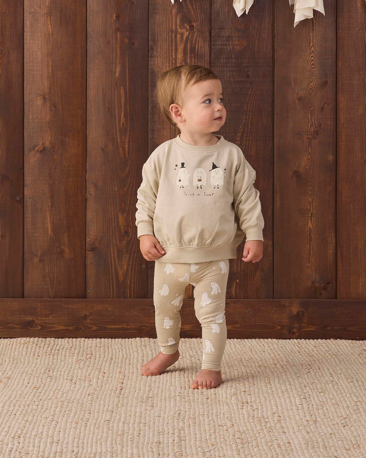 A toddler stands on a textured beige rug in front of a wooden wall, wearing the quincy mae relaxed fleece sweatshirt in trick or treat—crafted from organic cotton with playful animal faces—and matching leggings with white animal patterns, looking up to the side.