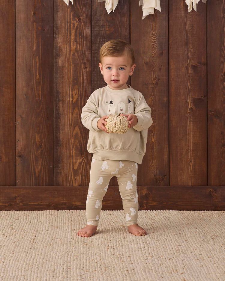 A toddler stands barefoot on a cream rug, wearing the quincy mae relaxed fleece sweatshirt in trick or treat, featuring playful trick or treat print and white ghost designs, holding a textured ball in front of a wooden wall with white decorations hanging above.