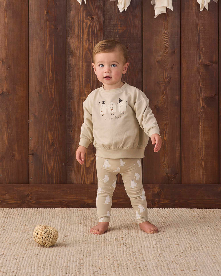 A toddler stands on a textured rug in front of a wooden wall, wearing the quincy mae relaxed fleece sweatshirt in trick or treat. A small decorative ball is on the floor nearby.