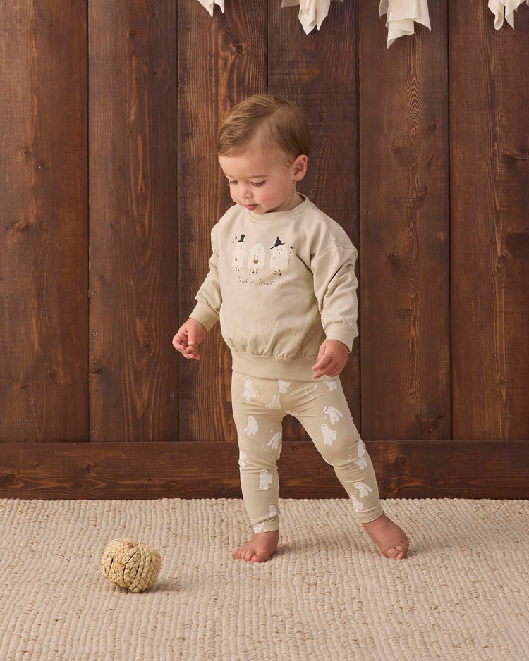A young child wearing the quincy mae relaxed fleece sweatshirt in trick or treat stands on a cream rug next to a textured ball, with a wooden wall in the background.