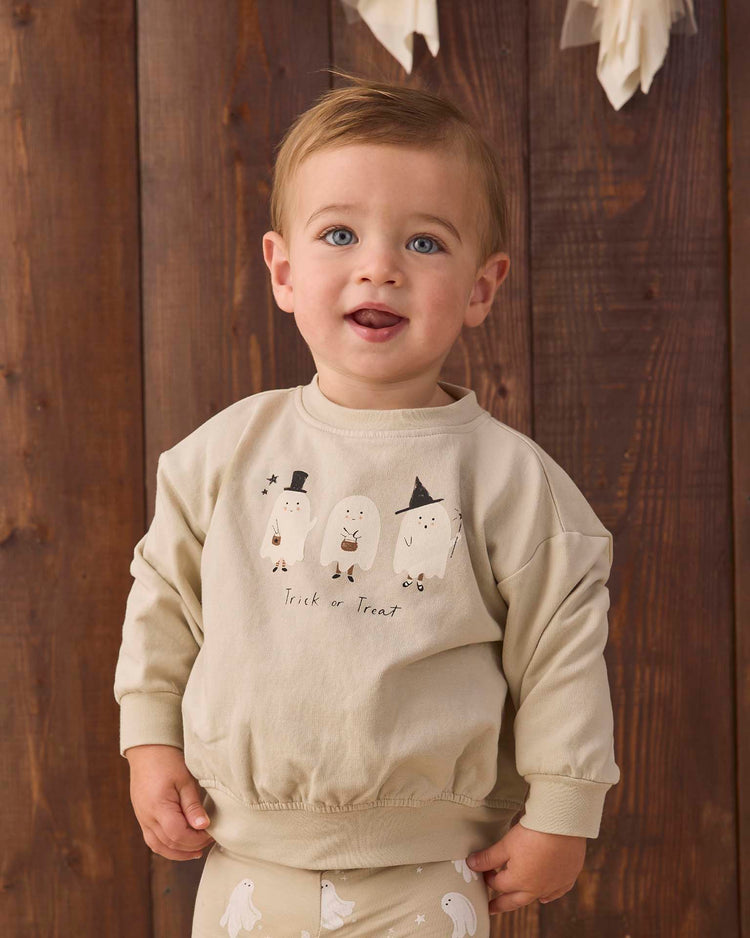 A toddler with light brown hair wears the quincy mae relaxed fleece sweatshirt in trick or treat, made from organic cotton and featuring playful ghosts and a fun trick or treat print, standing in front of a wooden background.