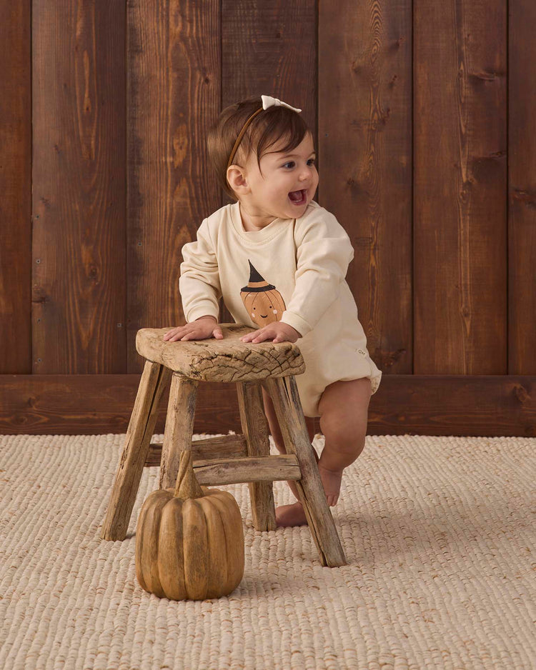 A smiling baby wearing the quincy mae crewneck bubble romper in lil pumpkin stands holding a rustic wooden stool, with a small pumpkin on the floor. The background features wood paneling and a woven rug.