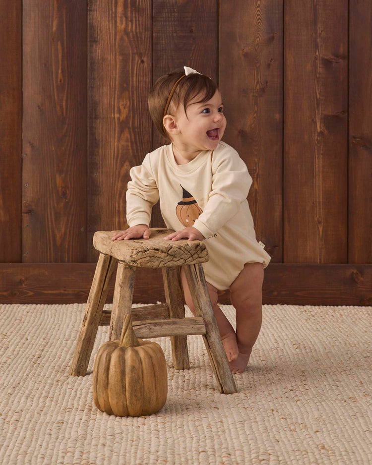 A smiling baby stands holding a rustic wooden stool, dressed in the quincy mae crewneck bubble romper in lil pumpkin. A decorative pumpkin sits on the floor in front, with wooden paneling in the background.