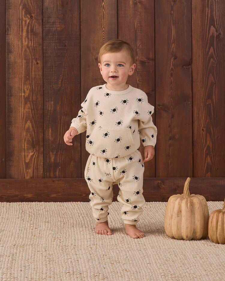 A toddler stands barefoot on a textured rug, wearing the quincy mae waffle slouch set in bitsy spider—a cream organic cotton ensemble decorated with black spider prints. Two light-colored pumpkins rest nearby against a wooden wall.