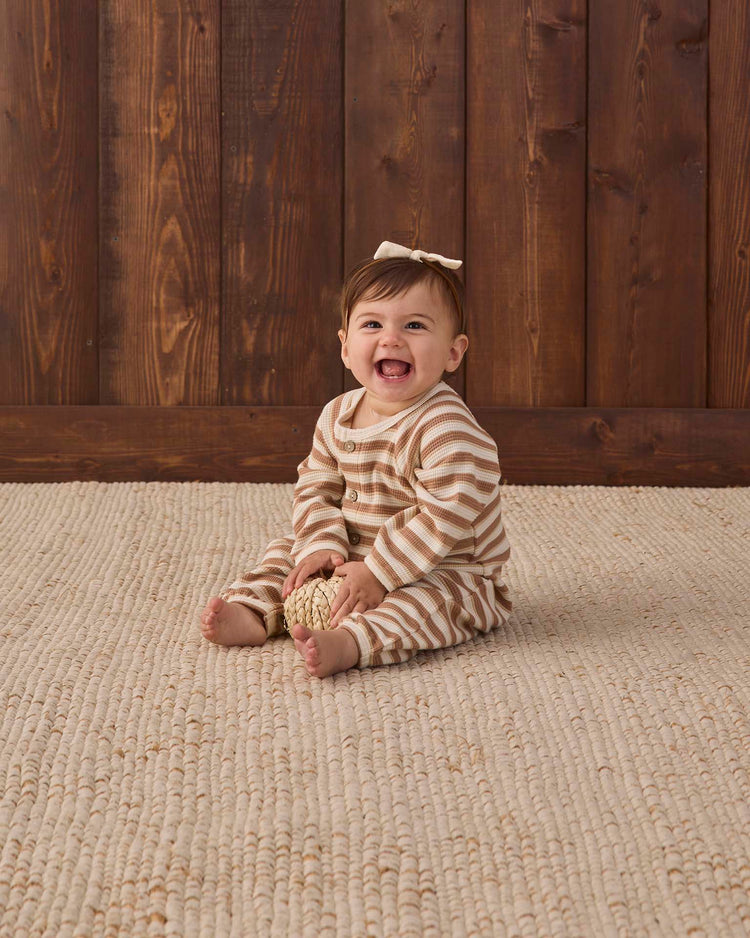 A smiling baby in the Quincy Mae waffle long sleeve jumpsuit in spice stripe, featuring adorable coconut buttons and a sweet bow, sits on a textured beige rug holding a small decorative ball, with a wooden wall in the background.