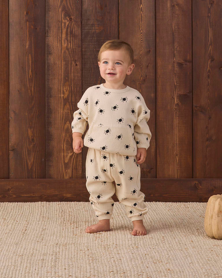 A toddler stands barefoot on a textured rug, wearing the quincy mae waffle slouch set in bitsy spider—a cream-colored kids loungewear set featuring a black spider print, crafted from soft organic cotton—in front of a wooden wall.