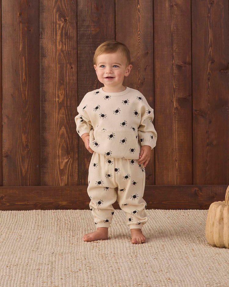 A toddler stands on a beige rug, wearing Quincy Mae’s waffle slouch set in bitsy spider—a cream-colored kids loungewear set adorned with playful black bug prints. Crafted from soft organic cotton, this outfit ensures maximum playtime comfort. Wooden panels form the background, with two small wooden pumpkins placed nearby on the floor.