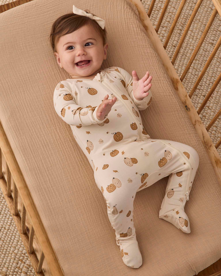 A smiling baby wearing the quincy mae bamboo zip footie in harvest friends—a light-colored outfit with a brown animal print—and a bow headband, lying on a beige mattress in a wooden crib.