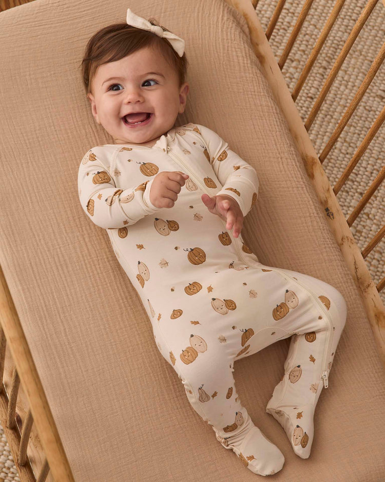 A smiling baby wearing the quincy mae bamboo zip footie in harvest friends, featuring pumpkin and gourd prints, lies on a beige blanket in a wooden crib and wears a matching bow headband.