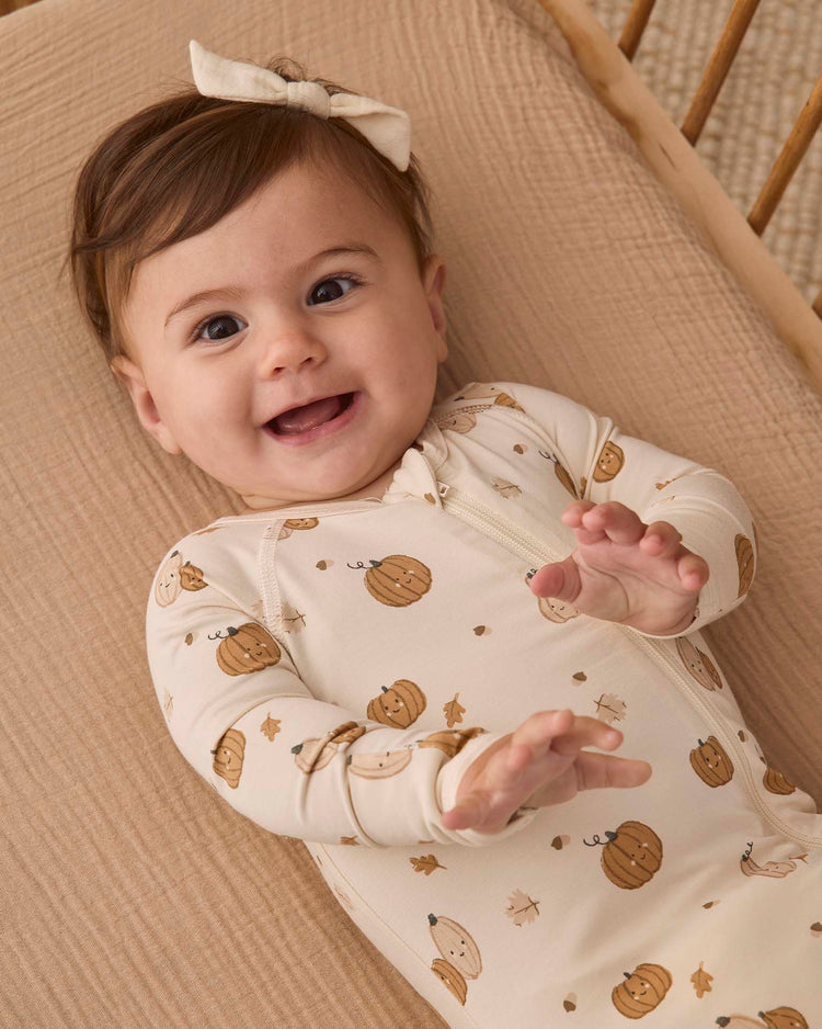 Smiling baby with brown hair and a bow, lying on a tan blanket, wearing the quincy mae bamboo zip footie in harvest friends print, reaching out with both hands.