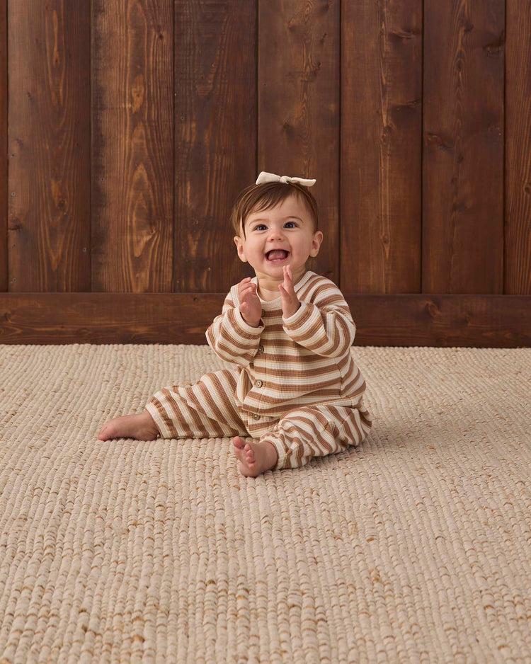 A smiling baby wearing the quincy mae waffle long sleeve jumpsuit in spice stripe, featuring coconut buttons and a bow on their head, sits clapping hands on a textured beige rug in front of a wooden wall.