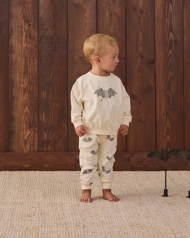 A young child stands on a textured rug in front of a wooden wall, wearing the quincy mae relaxed fleece sweatshirt in baby bats, featuring cream-colored fabric with bat motifs. A toy bat on a stand sits on the floor beside them.