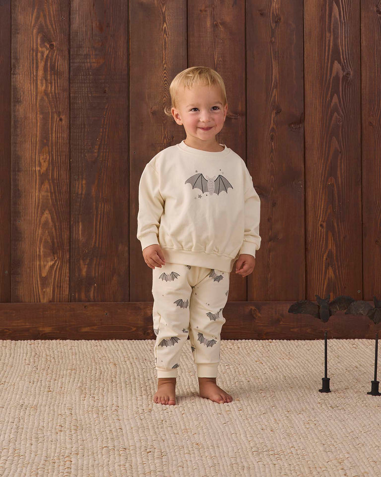 A toddler stands barefoot on a light rug, smiling in front of a wooden wall while wearing the quincy mae relaxed fleece sweatpant in baby bats, featuring a cream organic cotton fabric with gray bat print. Black bat decorations appear on the right.
