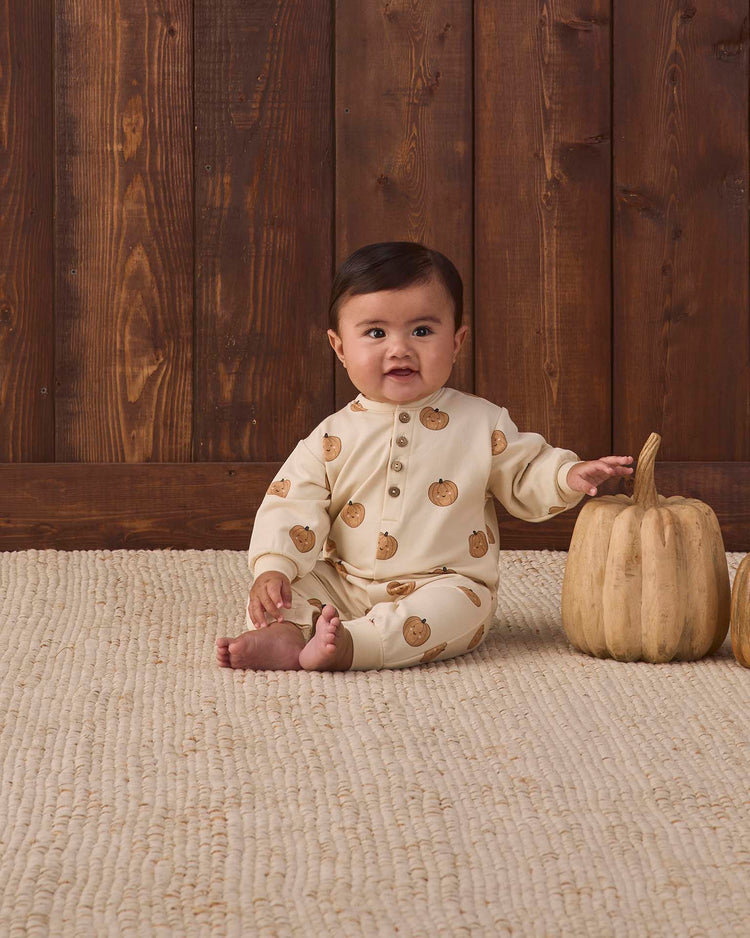 A baby wearing the quincy mae relaxed fleece jumpsuit in pumpkins sits on a textured rug beside a beige pumpkin, with a wooden wall in the background.