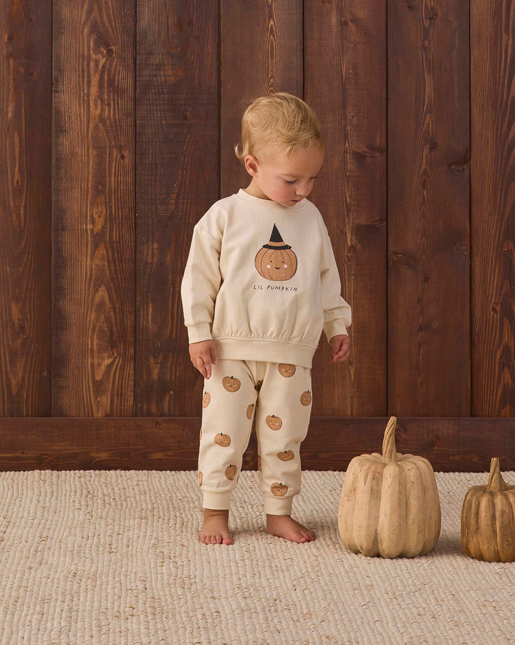 A toddler stands on a cream rug, wearing the quincy mae relaxed fleece sweatshirt in lil pumpkin. Two decorative pumpkins sit beside the child, while a wooden wall in the background adds a warm, rustic feel.