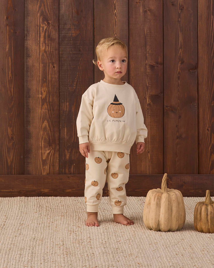 A toddler wearing the Quincy Mae relaxed fleece sweatshirt in lil pumpkin stands indoors on a tan rug, next to two beige pumpkins, in front of a wooden wall—creating the perfect cozy essential for autumn moments.