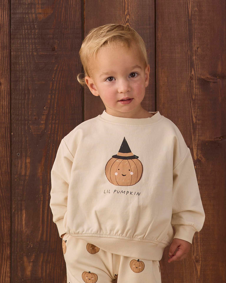 A young child stands in front of wooden boards, wearing the quincy mae relaxed fleece sweatshirt in lil pumpkin—a cozy cream organic cotton top featuring a smiling pumpkin graphic, a black hat, and the words “LIL PUMPKIN.” The look is completed with matching pants adorned with small pumpkin prints.