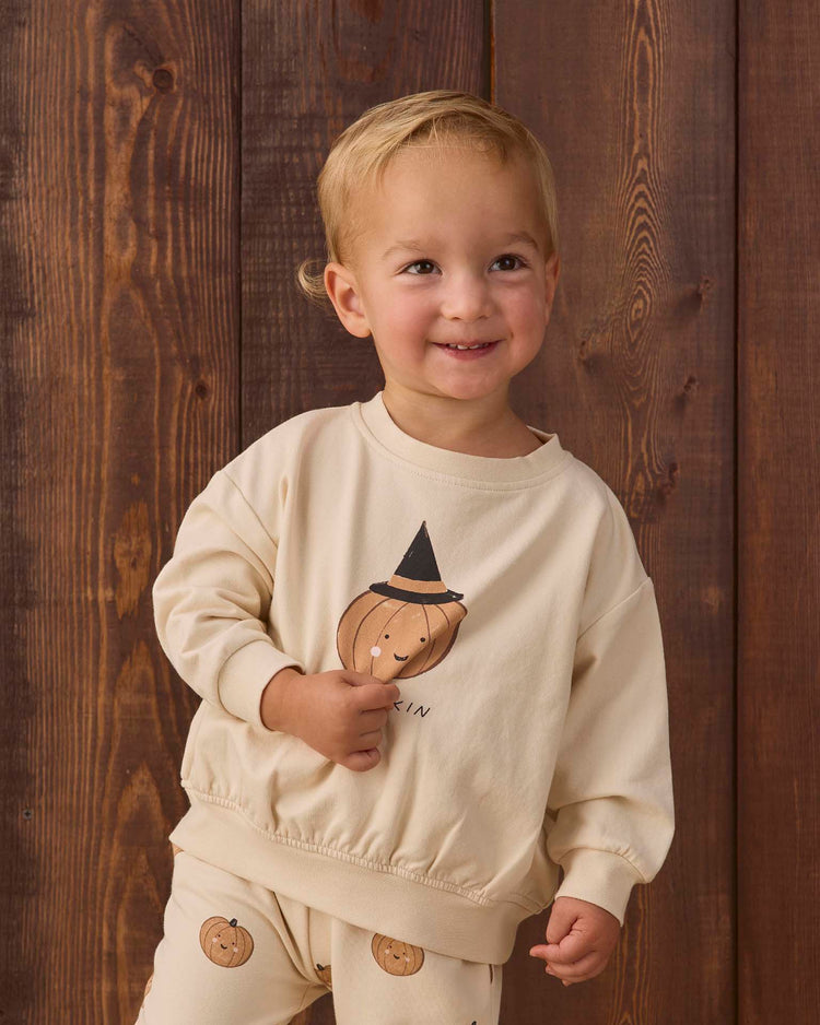 A smiling toddler wears the quincy mae relaxed fleece sweatshirt in lil pumpkin—an essential, cream-colored organic cotton top featuring playful pumpkin faces and a witch hat design—while standing in front of a wooden background.