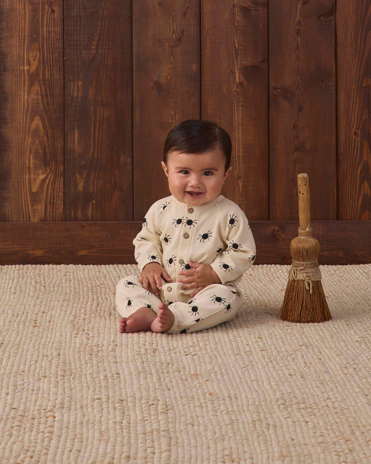 A baby wearing the quincy mae waffle long sleeve jumpsuit in bitsy spider print sits on a textured rug next to a small straw broom, with a wooden wall in the background.