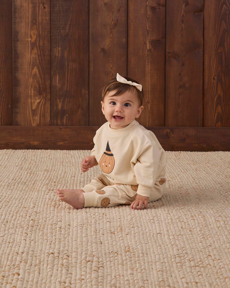 A smiling baby wearing quincy mae’s relaxed fleece sweatpant in pumpkins and a bow headband sits on a textured cream rug against a wooden wall background.