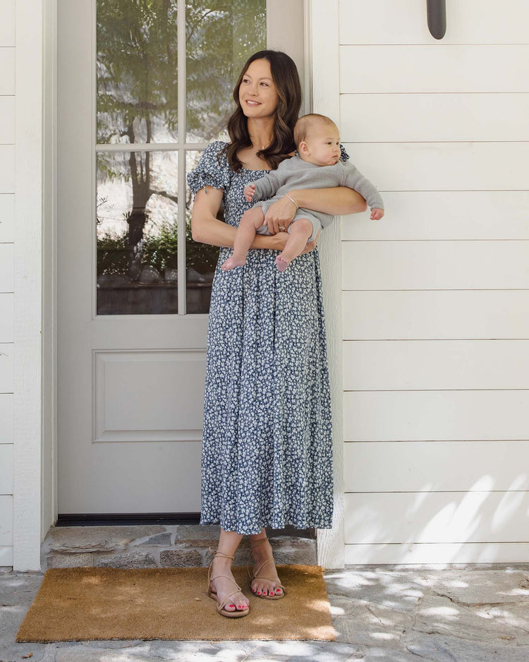A woman in a blue floral dress stands on a doormat before a white door, holding a baby dressed in the quincy mae summer knit set in heathered sky. They are outside with sunlight casting soft shadows on the ground.