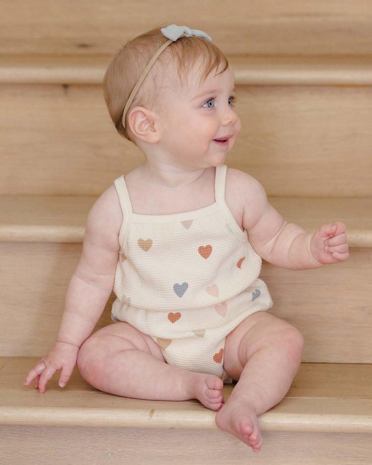 A smiling baby with light hair and a gray headband sits on wooden stairs, wearing the quincy mae waffle cinch romper in confetti hearts. The cream-colored organic cotton outfit features multicolored heart prints.