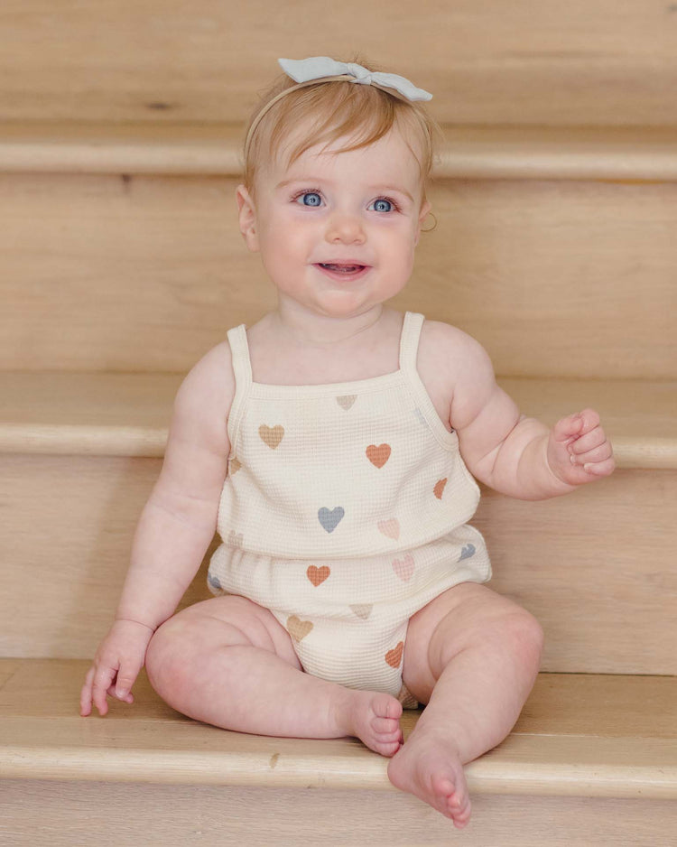 A smiling, light-haired, blue-eyed baby sits on wooden stairs wearing the quincy mae waffle cinch romper in confetti hearts and a light blue bow headband.