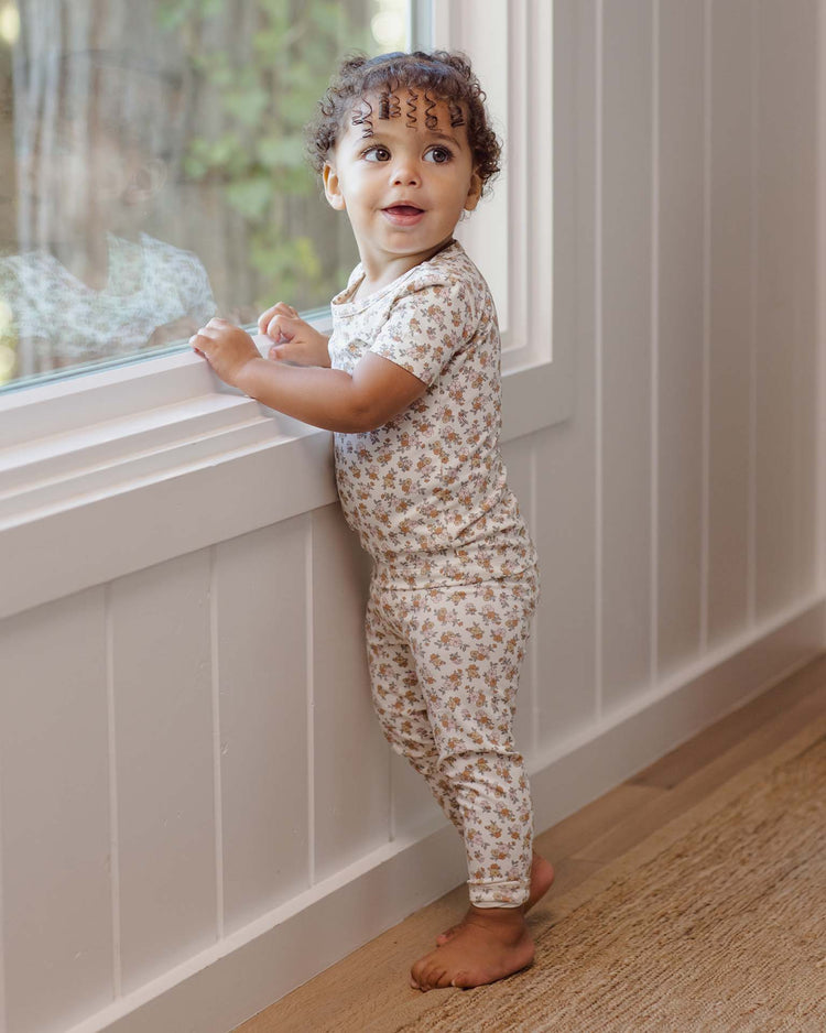 A toddler in the quincy mae bamboo short sleeve pajama set in wildflower stands barefoot by a sunny window, gazing upward with curiosity as their reflection appears in the glass.