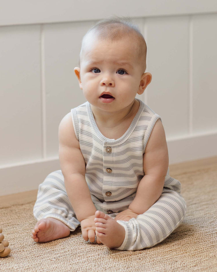 A baby wearing the quincy mae waffle jumpsuit in sky stripe sits on a woven rug, looking up with curiosity. The organic cotton fabric and soft blue and white stripes make it ideal for any transitional baby wardrobe.