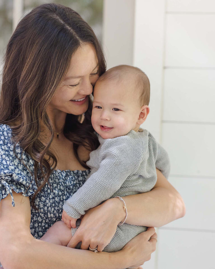 A woman with long brown hair in a blue floral dress smiles while holding a baby dressed in the quincy mae summer knit set in heathered sky, both looking happy and content in a bright indoor setting.