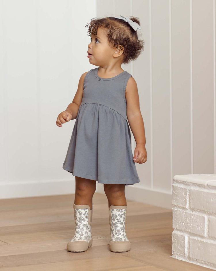 A young child with curly hair, wearing the quincy mae ribbed tank dress in ocean, stands indoors on a wooden floor near a white painted brick structure.