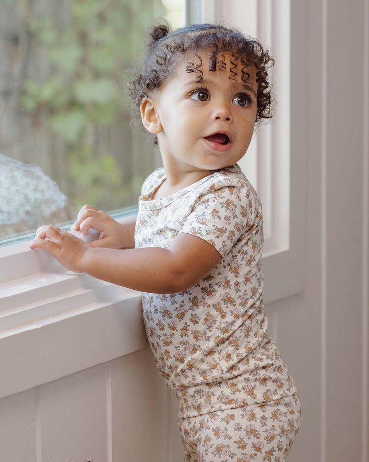 A toddler with curly hair wearing the quincy mae bamboo short sleeve pajama set in wildflower stands by a window, gazing outside with curiosity as sunlight fills the room.