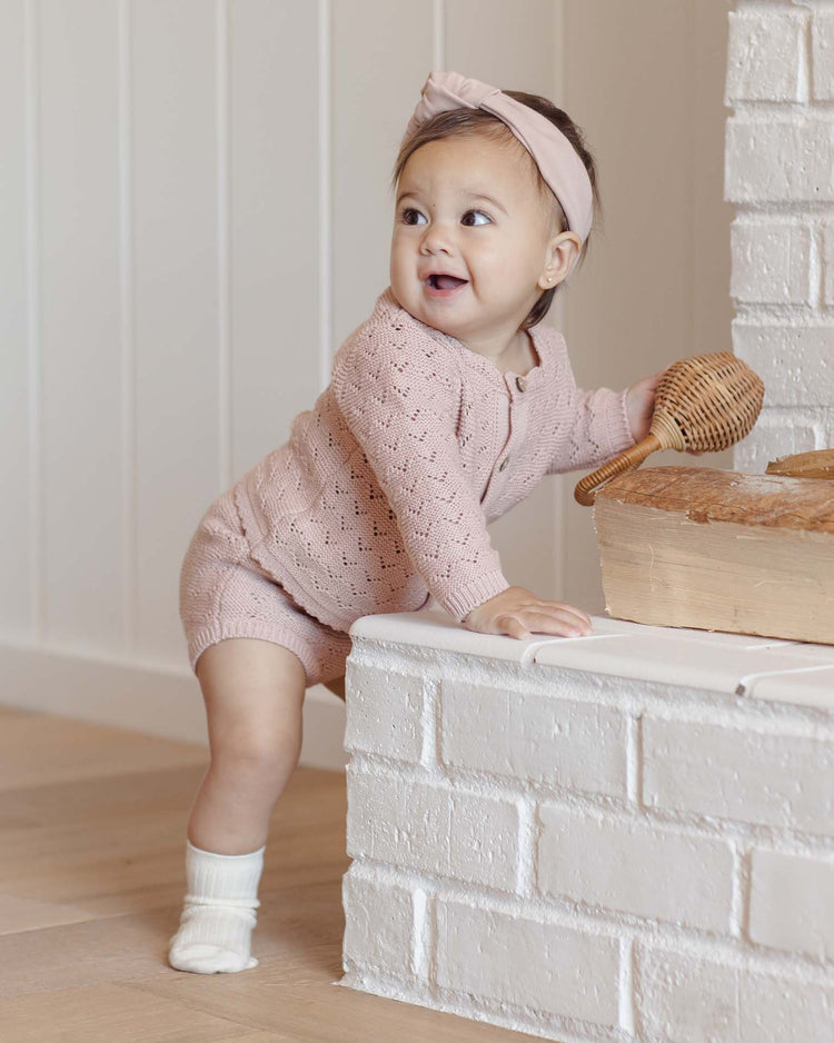 A baby in the quincy mae knit bloomer in blush stands by a white brick ledge, wearing white socks and a pink headband, holding a wicker rattle and smiling to the side.