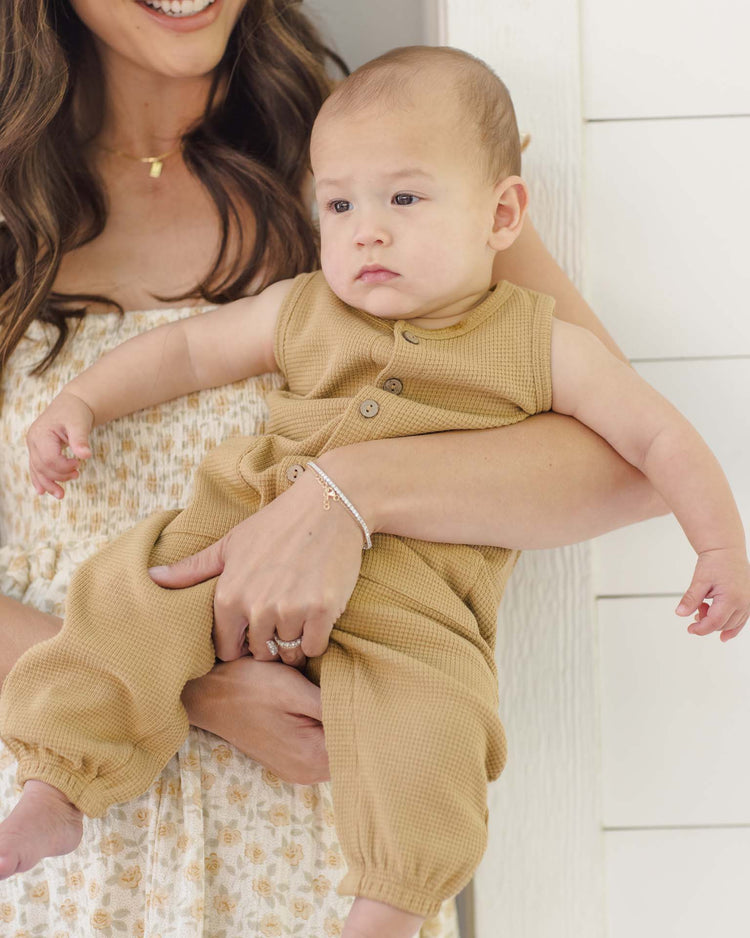 A woman in a floral dress holds a smiling baby wearing the quincy mae waffle jumpsuit in honey. They stand by a white wall, with the baby glancing to the side.