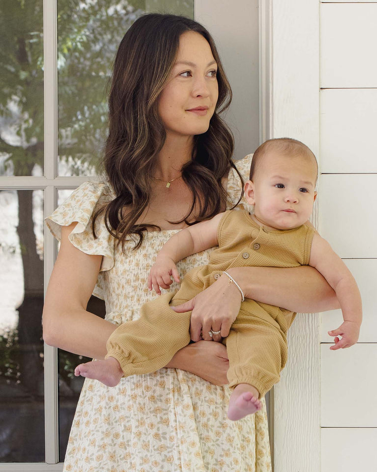A woman in a floral dress stands by a window, holding a baby wearing the quincy mae waffle jumpsuit in honey. The woman looks to the side while the baby faces forward, both calm as light streams in from outside.