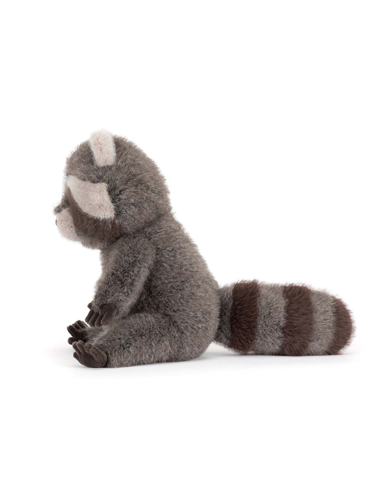 A jellycat trixiwoo raccoon stuffed animal sits upright, facing left, with gray fur, a striped tail, and white details on its face and ears against a plain white background.