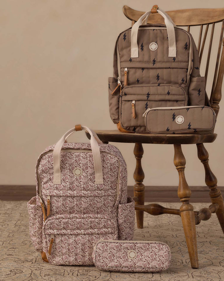 Two patterned backpacks and matching pencil pouches—essential school supplies—are displayed on a wooden chair and a textured rug. One set features a light pink backpack with floral designs; the other includes the beige rylee + cru pencil pouch in bolts paired with a backpack adorned with dark lightning bolt prints.