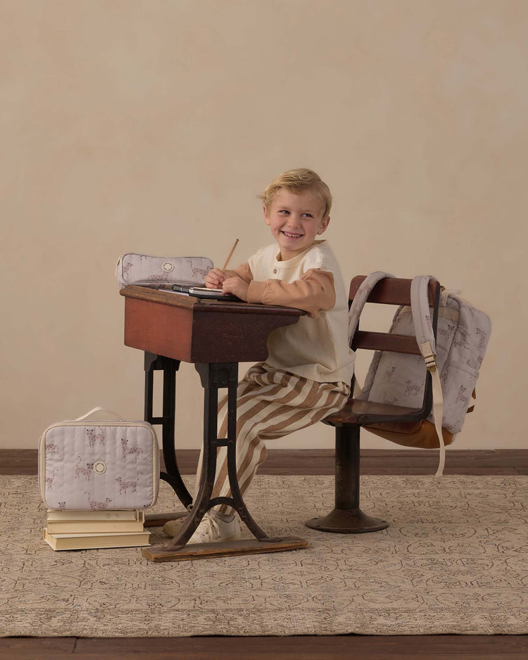 A young child sits at an old-fashioned wooden desk, smiling with a pencil in hand. Nearby are a backpack, a rylee + cru lunch bag in leopard print, and a pencil case. The child is dressed in striped pants and a cream top.