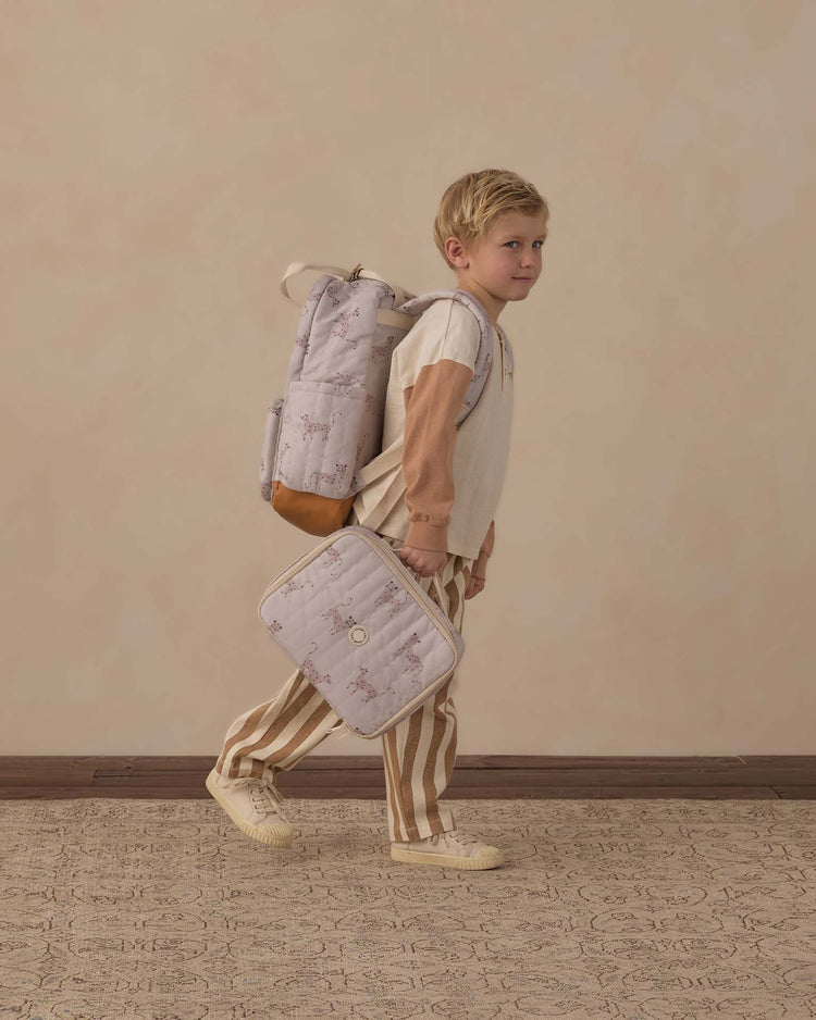 A young child with blond hair wears striped pants, a long-sleeve shirt, and sneakers, carrying the rylee + cru backpack in leopard along with a matching lunch bag—ready for everyday adventures while walking indoors against a beige wall.