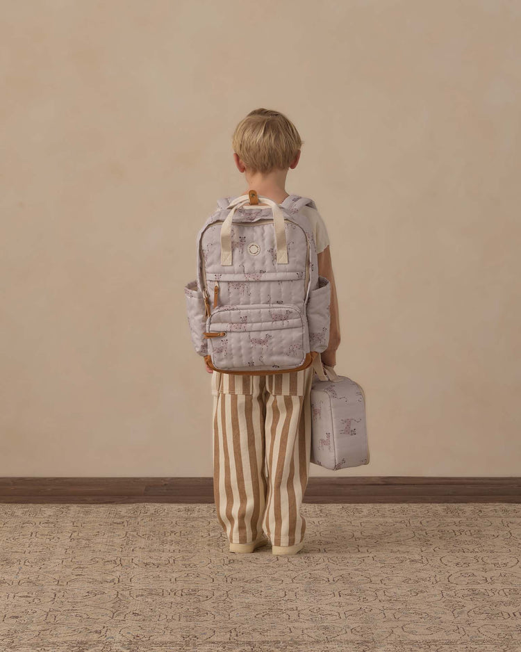 A child with short blond hair, wearing striped pants and a light shirt, stands facing a beige wall, holding a lunchbox in one hand and wearing the rylee + cru backpack in leopard—a light-colored bag crafted from durable polyester, perfect for everyday adventures.