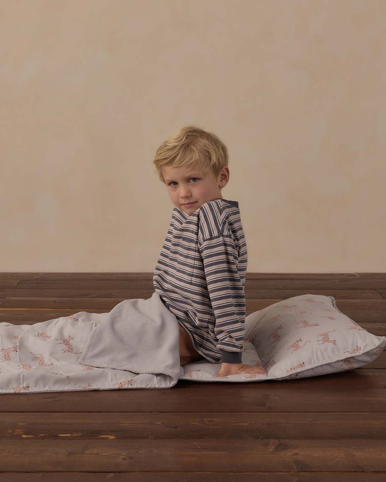 A young child in striped pajamas sits on the rylee + cru nap mat in leopard, which features a removable pillow and a soft micro-fleece blanket, on a wooden floor, looking toward the camera with a neutral expression.