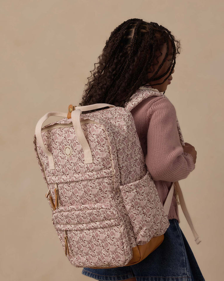 A young girl with braided hair wearing a mauve sweater and denim skirt stands sideways, carrying the rylee + cru backpack in primrose—a pink and white floral-patterned bag with tan accents, featuring a spacious main compartment perfect for all her everyday adventures.