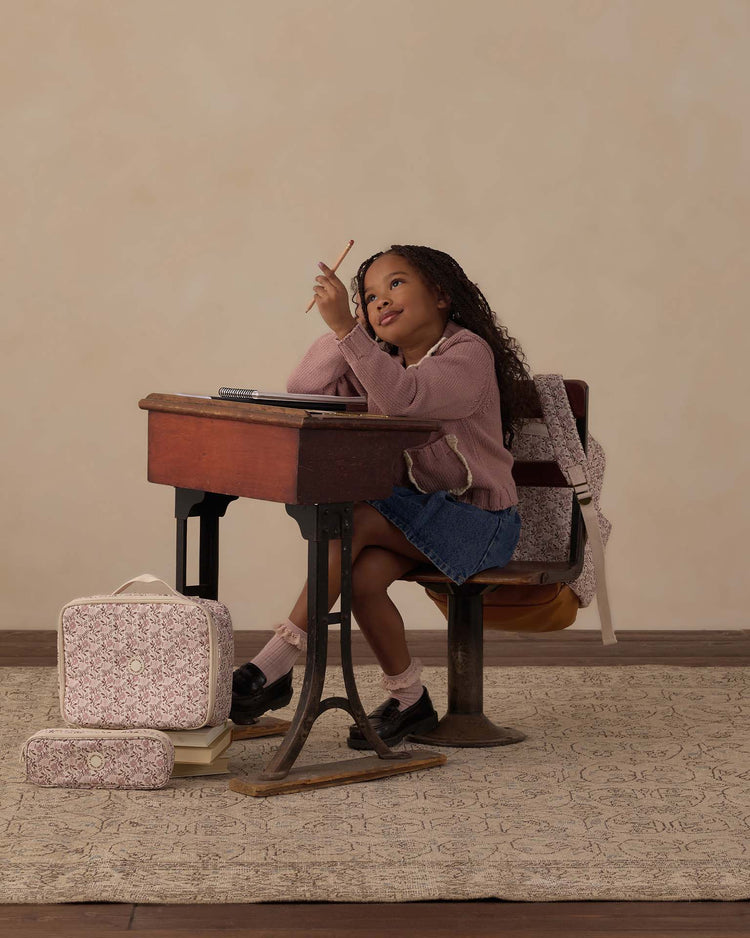 A young girl sits at a vintage school desk, smiling and looking up while holding a pencil. Beside her, she has a backpack and patterned bags—including the rylee + cru pencil pouch in primrose for her school essentials. She wears a pink sweater and denim skirt against a neutral, soft background.