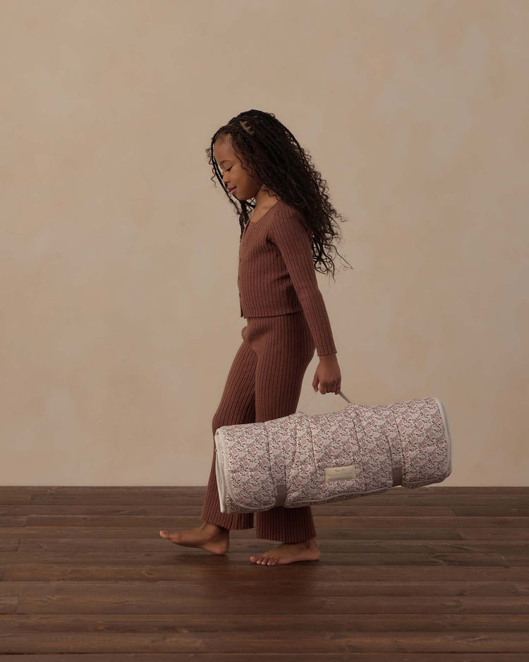 A young girl with long curly hair walks barefoot on a wooden floor, wearing a ribbed brown outfit and carrying the rylee + cru nap mat in primrose, rolled up in its fabric bag. The background is a plain beige wall.